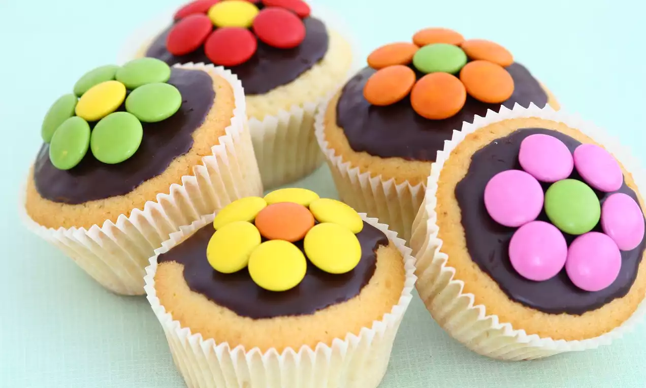 Vanilyalı muffinler açık mavi zeminde, parlak çikolata kaplaması ve çiçek şeklinde renkli draje süslemesi ile bir arada düzenlenmiş