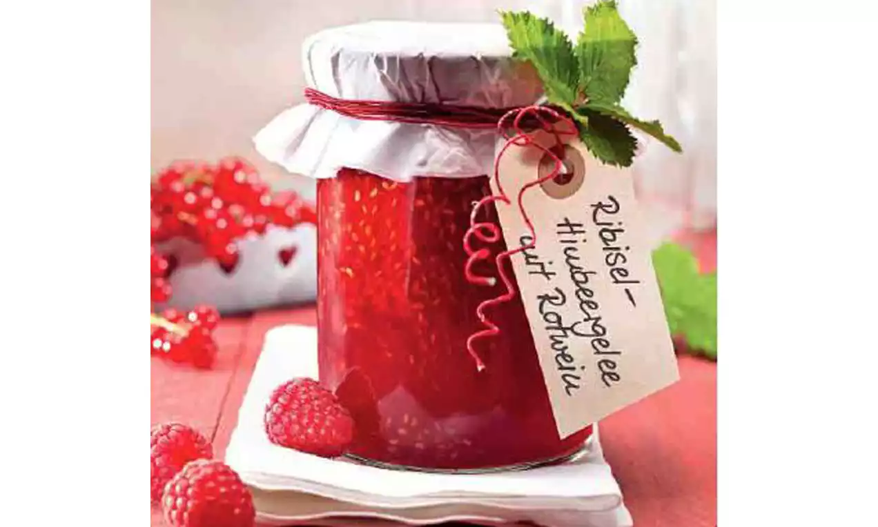 Ein Glas leuchtend rotes Gelee mit Ribiseln und Himbeeren, abgedeckt mit weißem Papierdeckel und rotem Band, dekoriert mit Etikett und grünen Blättern