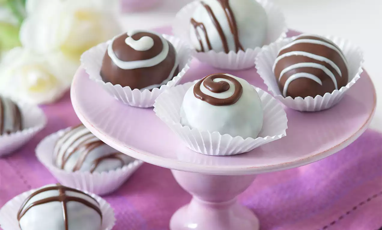 Čokoládové guľôčky v bielych a hnedých polevách so vzorom špirál a prúžkov, usporiadané v papierových košíčkoch na ružovom stojane.