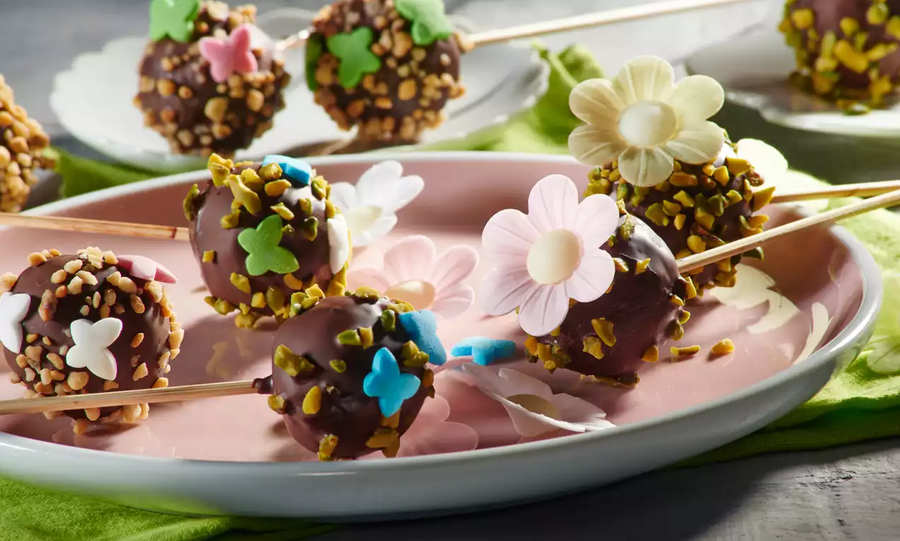 Schokoladige Cakepops mit bunten Tupfen aus Zuckerstreuseln und dekorativen Blüten auf Holzspießen auf einem rosa Teller mit grünem Stoff