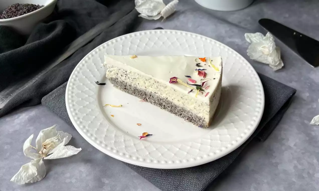 Makový cheesecake s vrstvou svetlého makového cesta a krémovo bielej plnky, ozdobený farebnými jedlými kvetmi na bielom tanieri.
