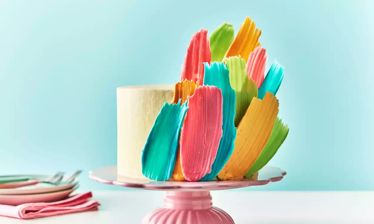 Tall white cake on a pink stand adorned with vibrant chocolate brushstrokes in blue, green, yellow, orange and pink against a pale blue backdrop