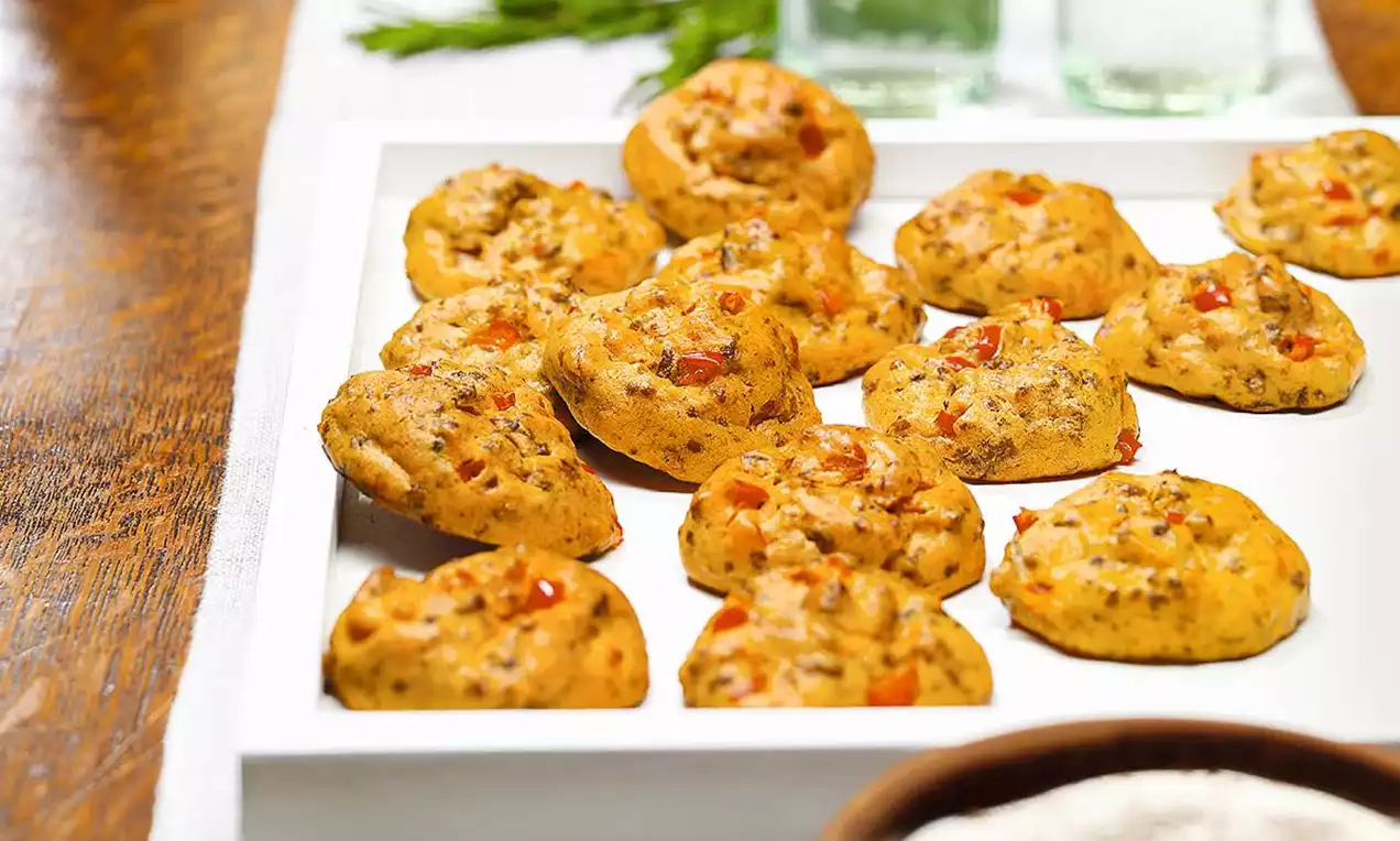 Malé okrúhle stredomorské pečivo zlatohnedého odtieňa, s kúskami zeleniny a mäsa, poukladané na bielom podnose s bylinkami v pozadí