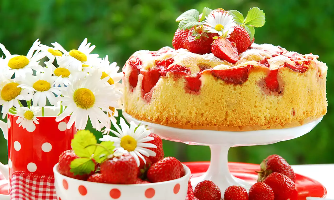 Pandișpan pufos auriu cu căpșuni roșii coapte, decorat cu zahăr pudră, flori albe și frunze verzi, așezat pe un platou alb