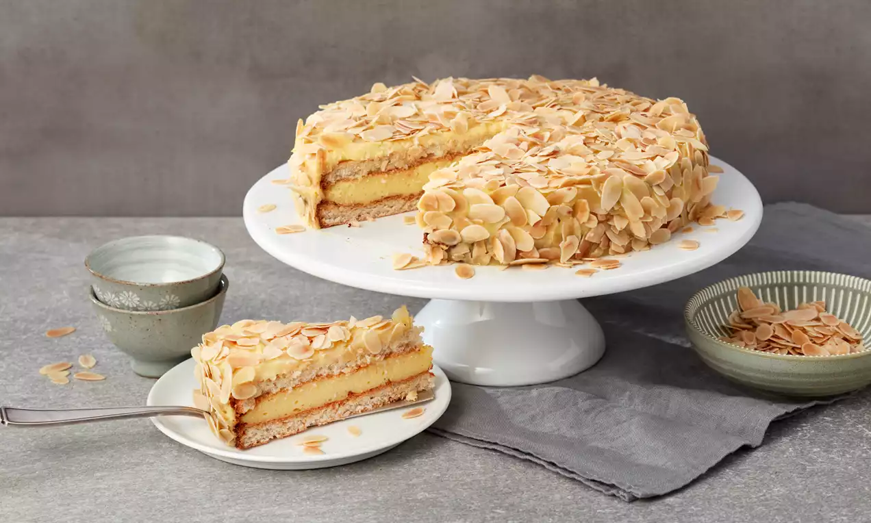 Schwedische Mandeltorte mit goldbraunen, gerösteten Mandelsplittern auf leichtem gelbem Teig, serviert auf weißer Tortenplatte mit Beilagenteller