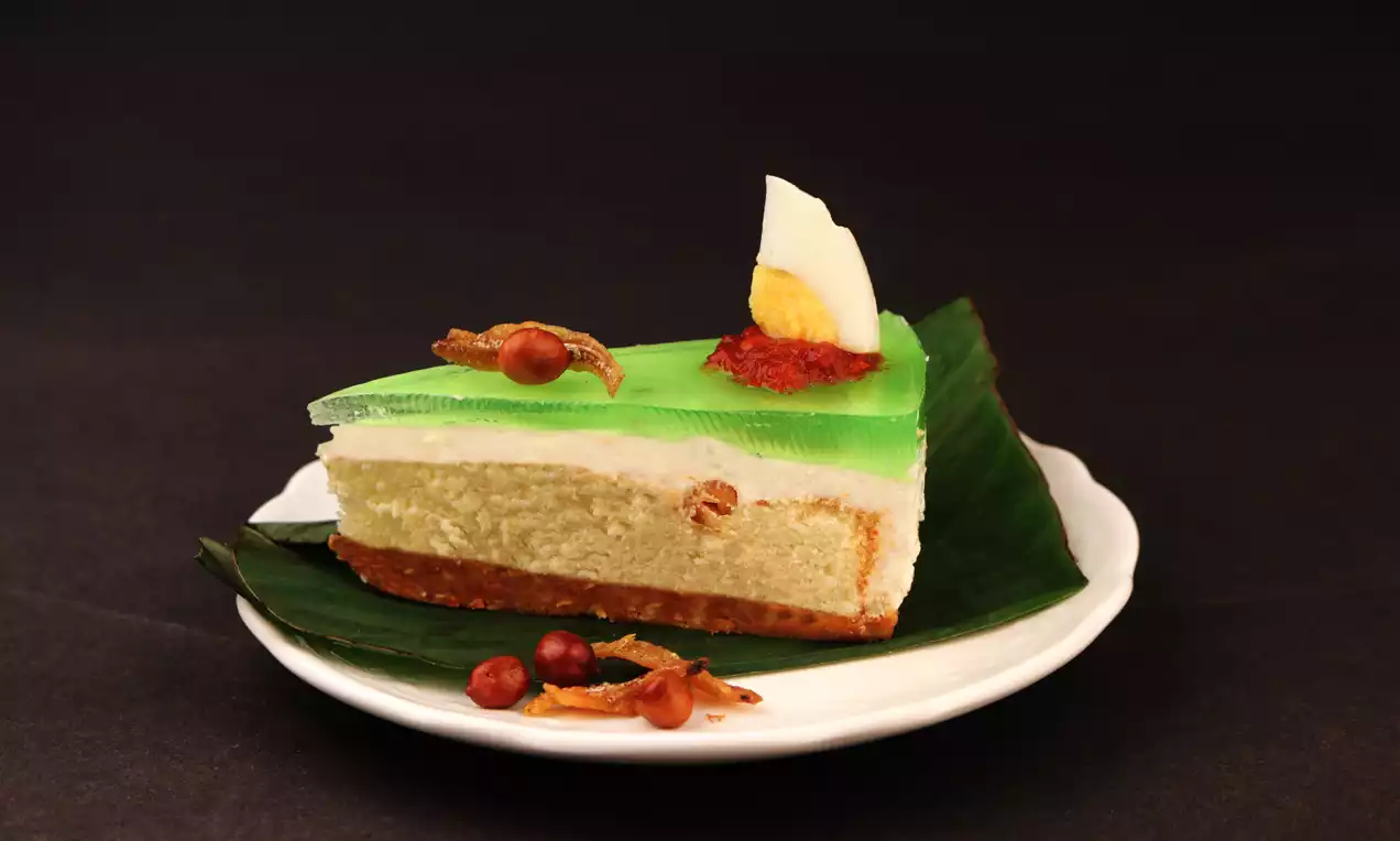 A slice of sponge cake with green jelly topping, white cream layer, garnished with half-boiled egg, sambal, peanuts and anchovies on banana leaf