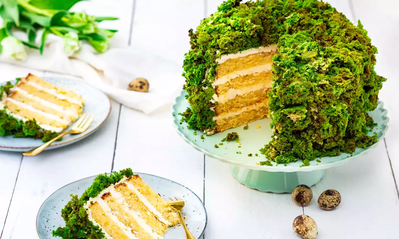 Tort verde vibrant cu blat stratificat galben-auriu și cremă albă, decorat cu mușchi de copac pe un suport pastel pe masă albă