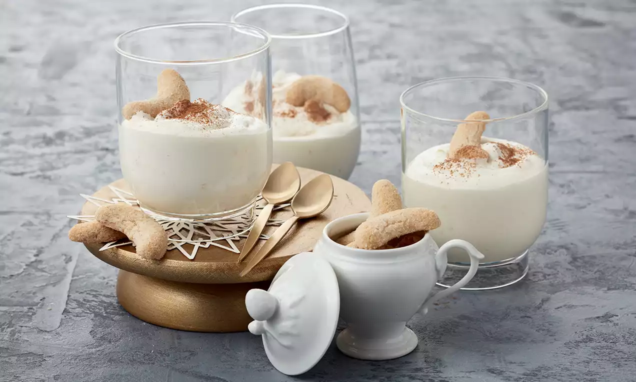 Glas mit cremig weißer Vanillekipferlcreme, bestäubt mit braunem Zimt, garniert mit goldbraunem Kipferl, auf Holzplatte mit Dekor drapiert