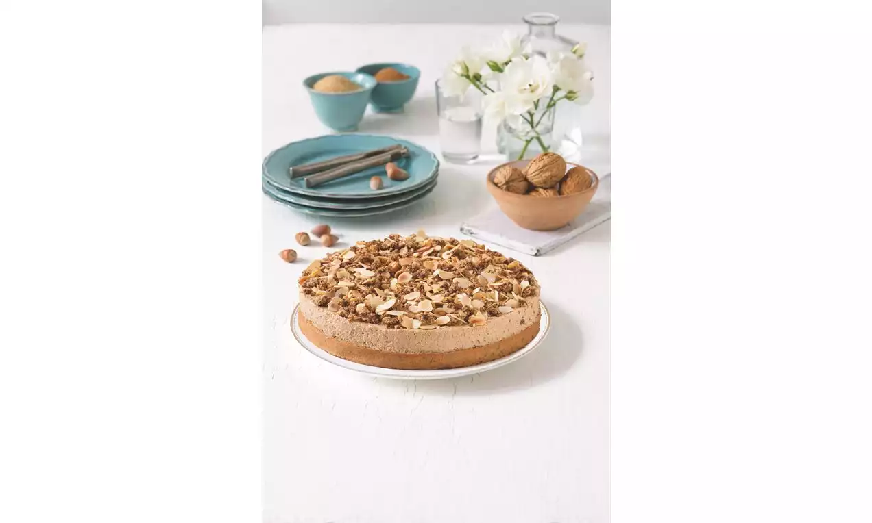 Podzimní oříškový koláč se světlým nadýchaným krémem na zlatavém základu, posypaný směsí lískových a vlašských ořechů na bílém talíři