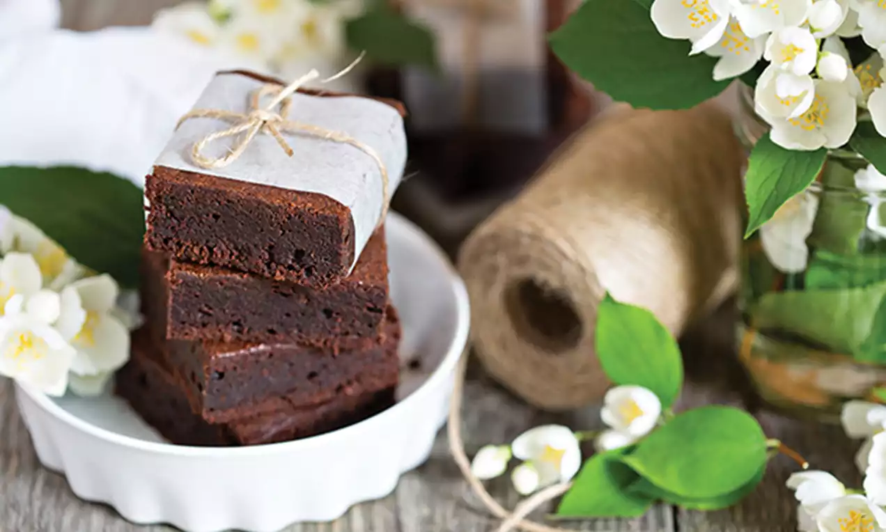 Stacked chocolate brownies with rich, dark interiors wrapped in parchment and twine, on a white dish surrounded by jasmine flowers