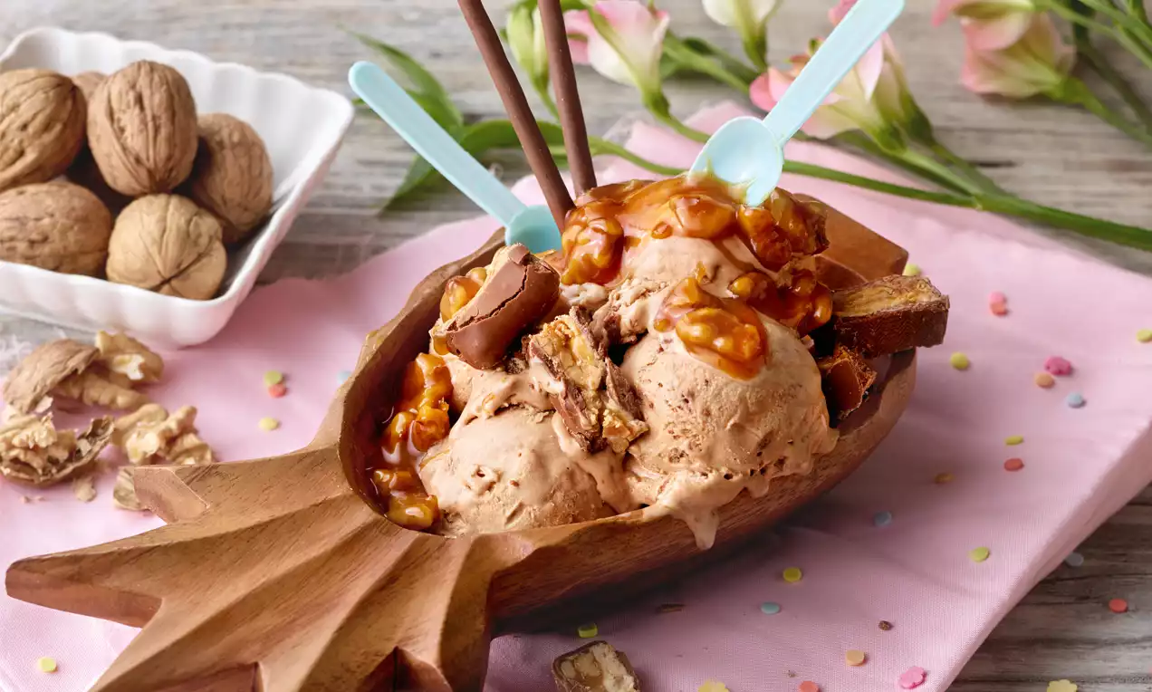 Cremiges Walnusseis in einem braunen Holzschalenbecher, dekoriert mit Karamellsauce, Schokostäbchen, Walnüssen und bunten Streuseln
