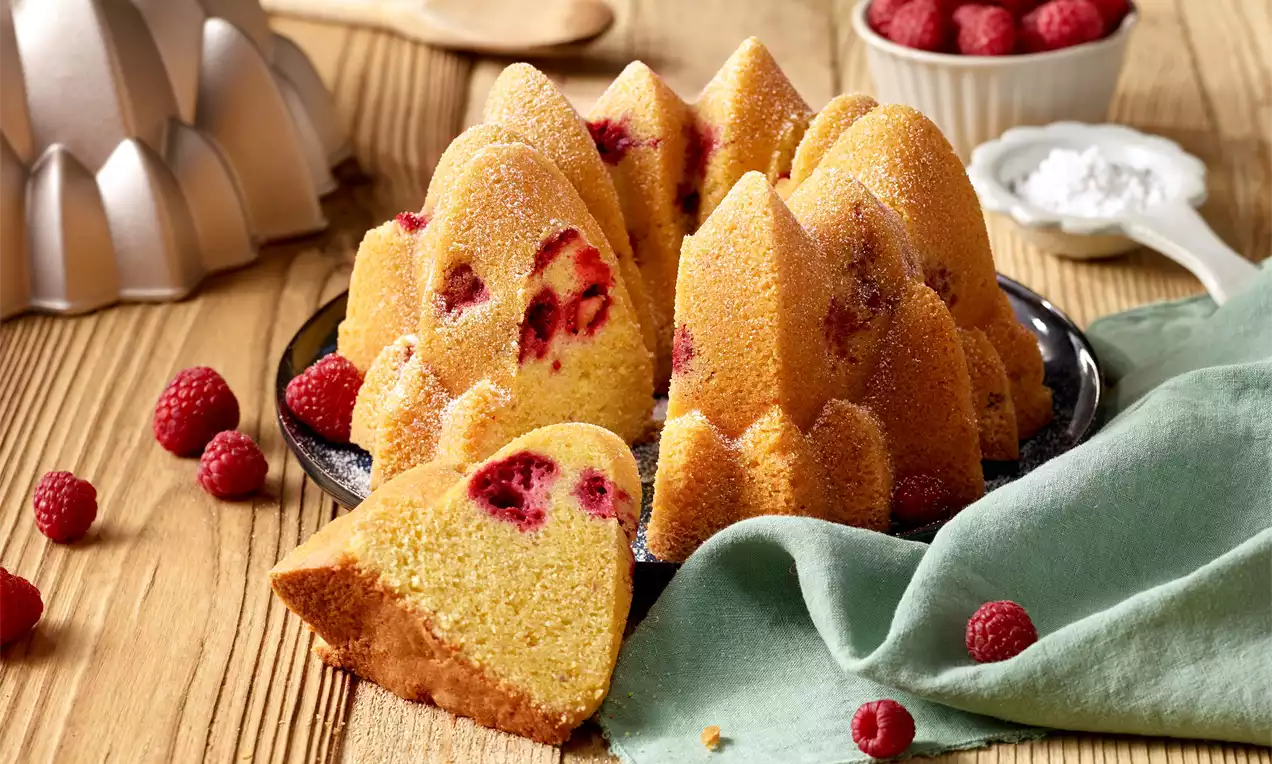 Zlatistá bábovka v tvare vysokých oblúčikov s červenými malinami vo vnútri, posadená na tmavom tanieri, obklopená čerstvými malinami a zelenou látkou