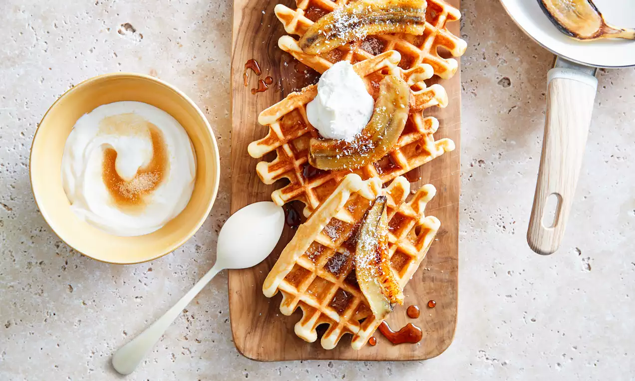 Gaufres dorées aux bords croquants garnies de bananes caramélisées, crème blanche et filet de sirop sur une planche en bois