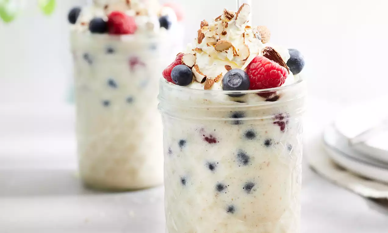 Veganski shake od vanilije u staklenoj čaši s uzorkom, kremaste teksture s borovnicama i malinama, ukrašen šlagom, čokoladnim mrvicama i lješnjacima