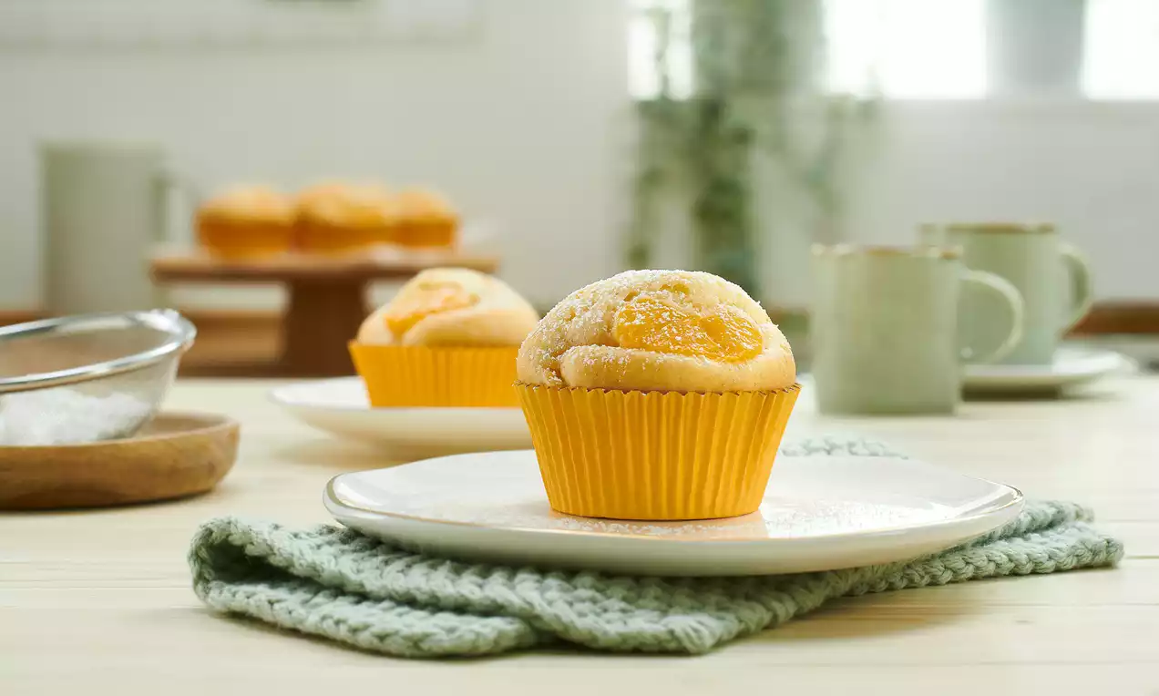 Ein goldgelber Mandarinen-Muffin in orangefarbener Papierform auf weißem Teller mit grünem Strickuntersetzer in heller Tischszenerie