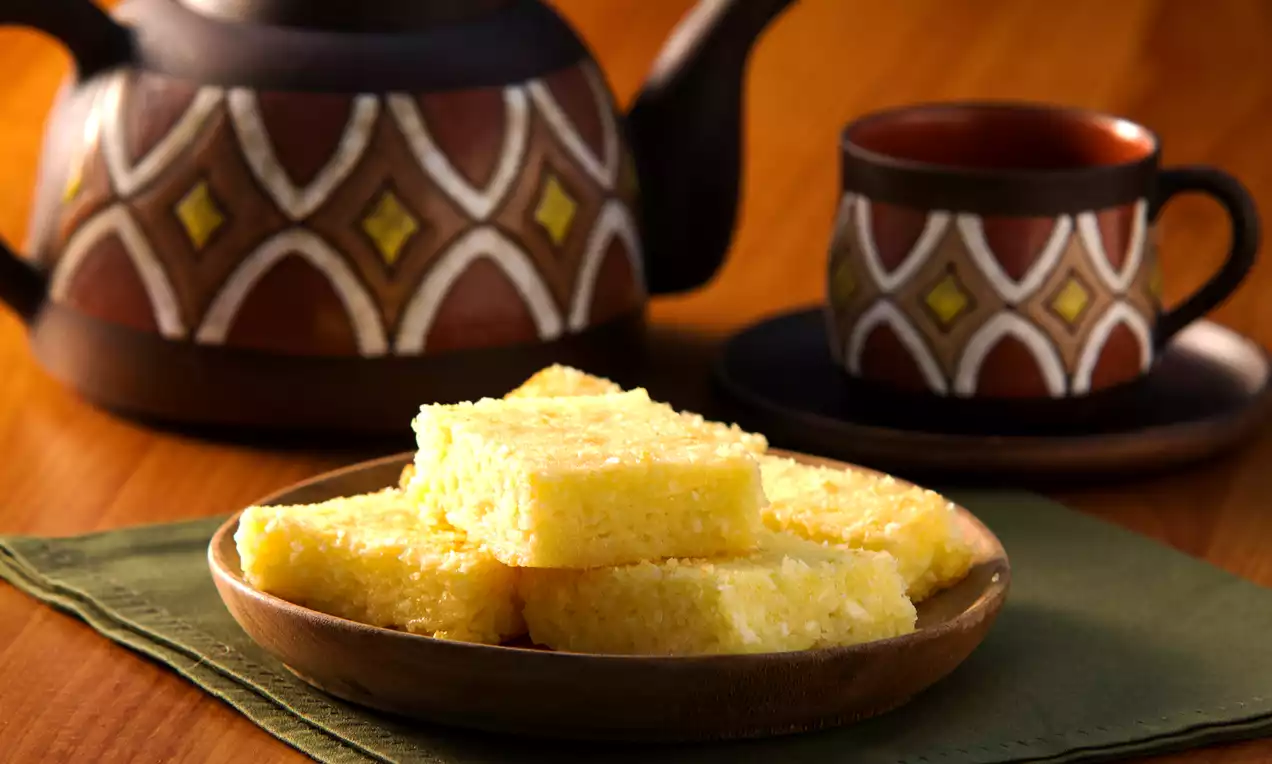 Pedaços retangulares de bolo de mandioca amarelo claro com textura macia em prato rústico sobre guardanapo verde, ao lado de bule e xícara decorados