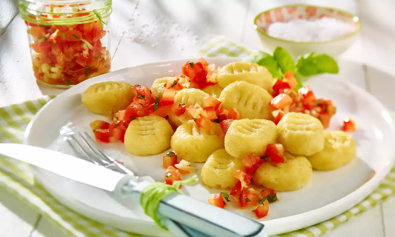 Goldgelbe Gnocchi mit geriffelter Oberfläche, bedeckt von feiner, rot-grüner Tomatensalsa, auf weißem Teller mit Basilikumblättern