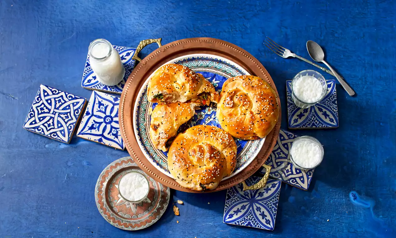 Goldbraune Börek mit Spinatfüllung, sesam-bestreut auf gemustertem Teller, arrangiert mit kleinen Gläsern Joghurt auf blauem Untergrund
