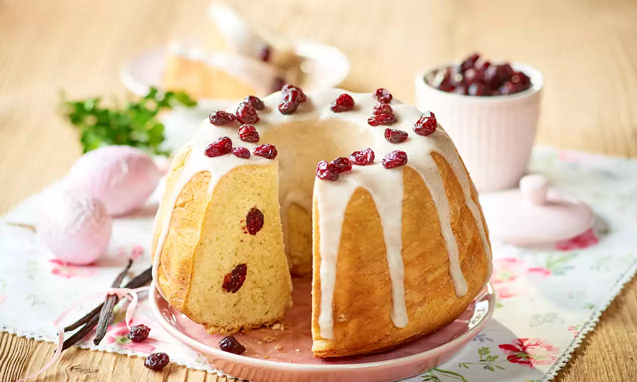 Waniliowa babka drożdżowa o złocistej skórce z białą polewą lukrową, udekorowana suszoną żurawiną, na różowym talerzu na kwiecistym obrusie