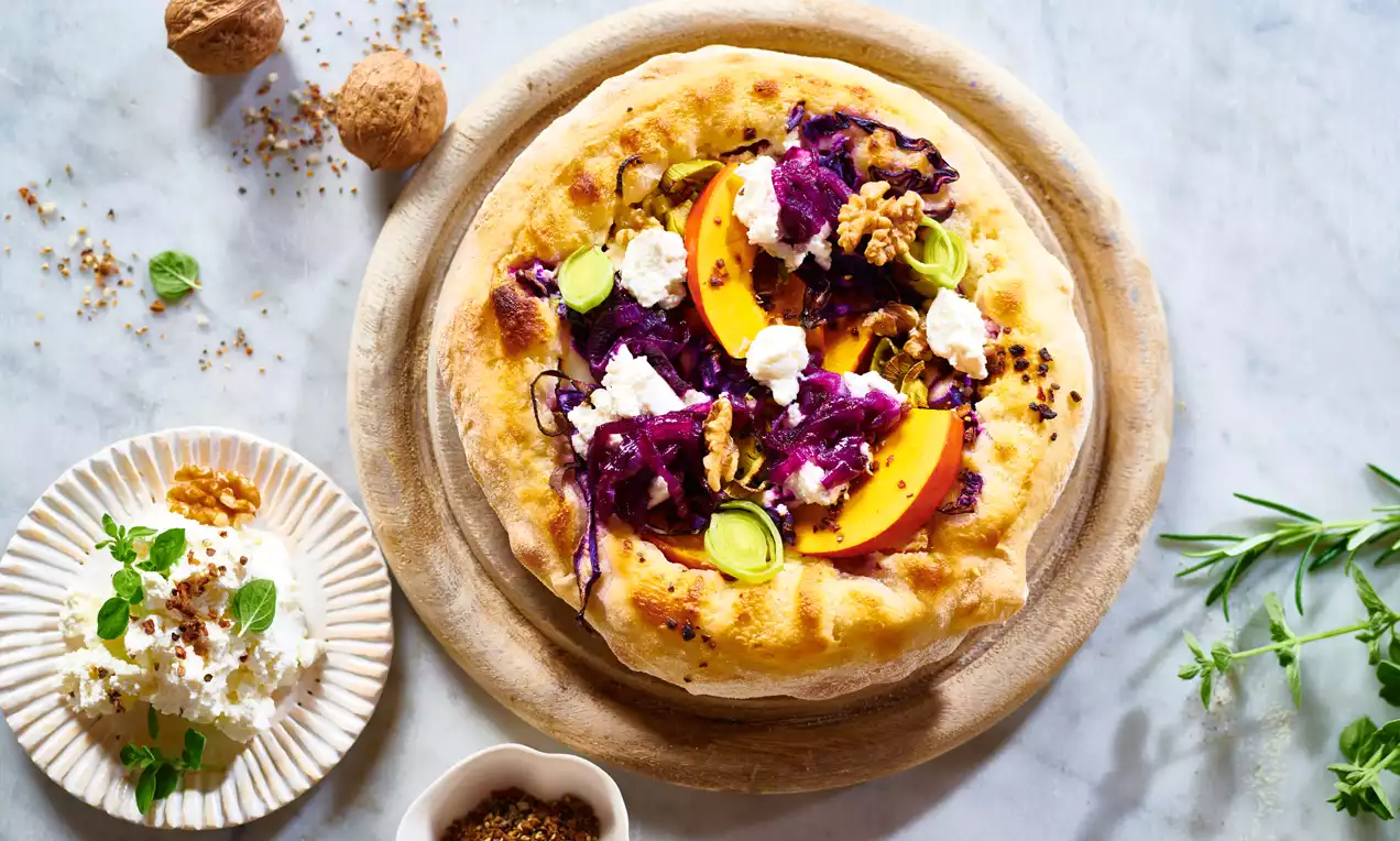 Winter-Pizza mit goldbraunem Rand, belegt mit Kürbisspalten, Rotkohl, Walnüssen, Ziegenkäse und Zucchini auf hellem Holzbrett