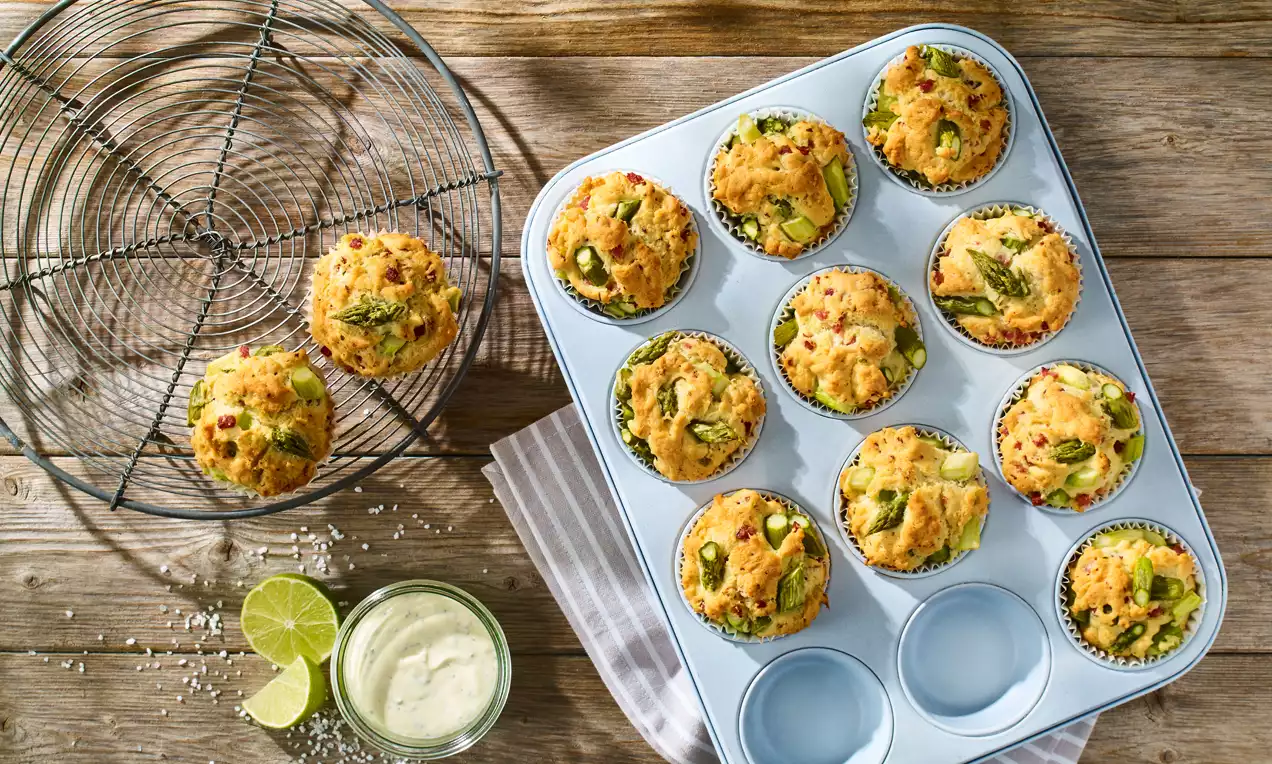Spargelmuffins mit grünem Spargel und Schinkenwürfeln, goldbraun gebacken, auf einem Holzbrett mit kleinem weißen Dip
