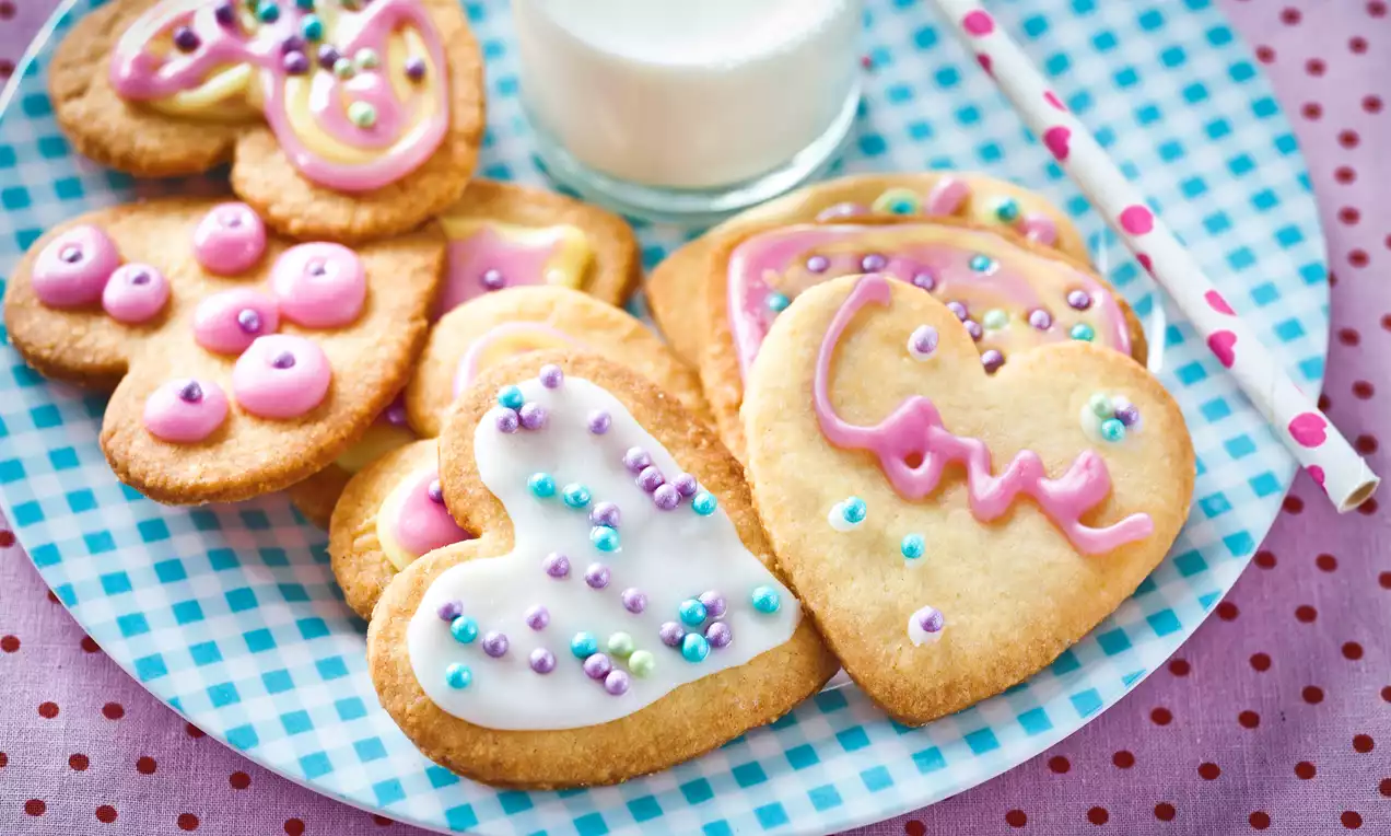 Kruche ciasteczka w kształcie serc, ozdobione kolorowym lukrem, pastelowymi perełkami i ułożone na niebieskim talerzu w kratkę