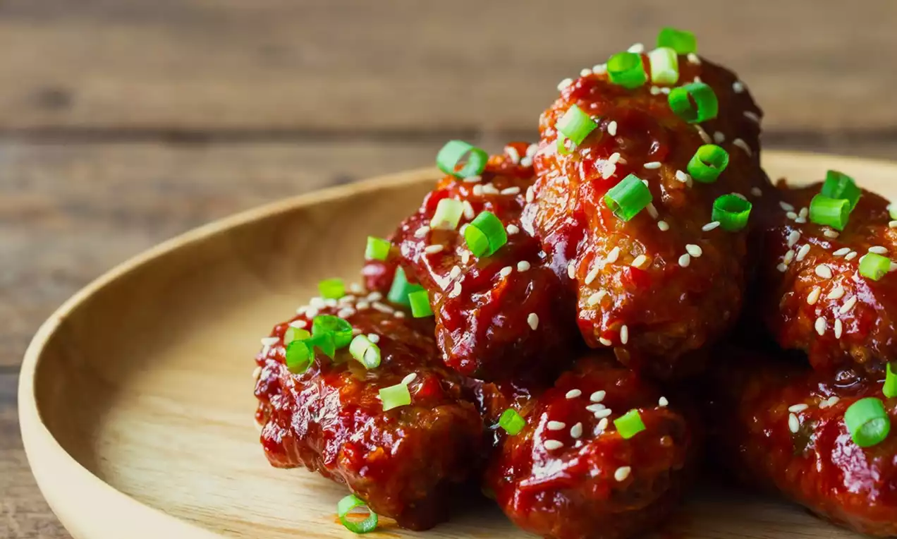 Crispy Korean fried chicken pieces glazed in shiny red sauce, topped with white sesame seeds and green onion slices on a wooden plate