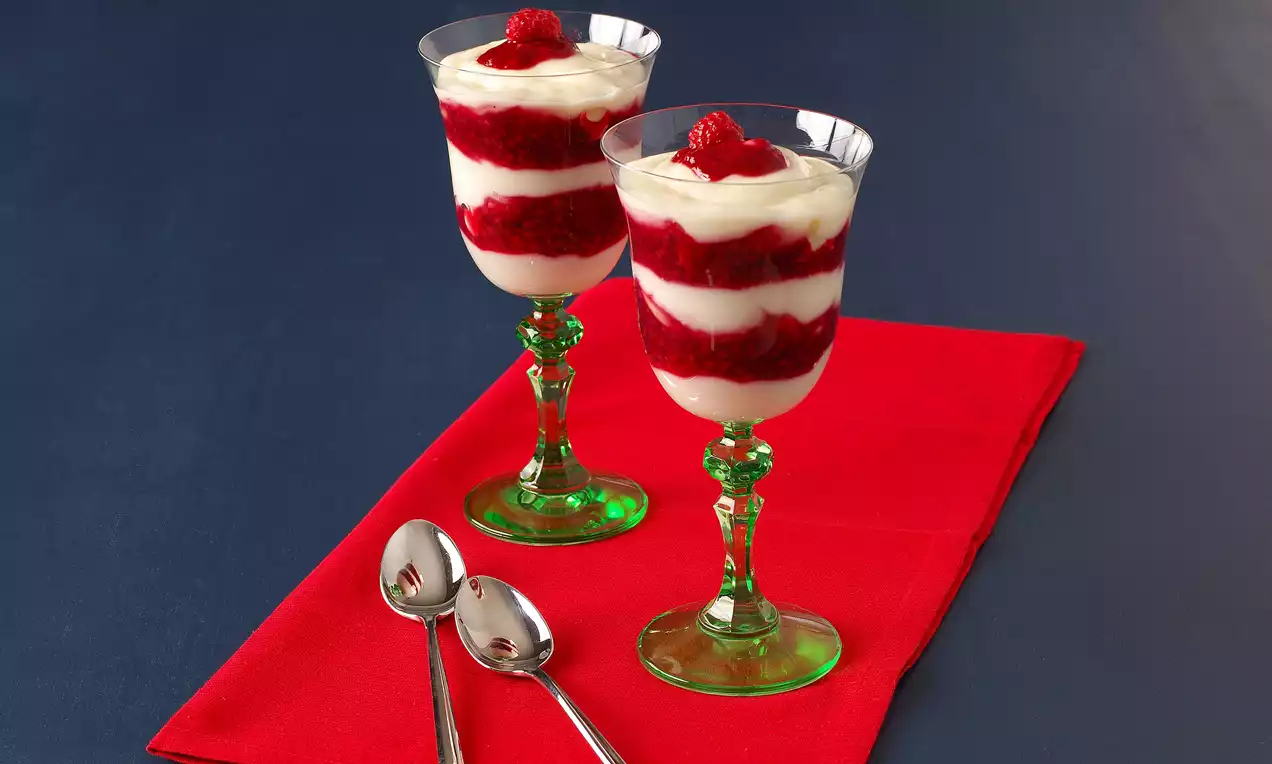 Yeşil ayaklı şık kadehlerde kat kat ahududu püresi ve beyaz sakızlı muhallebi, üstünde taze ahududu ile kırmızı bir örtü üzerinde 