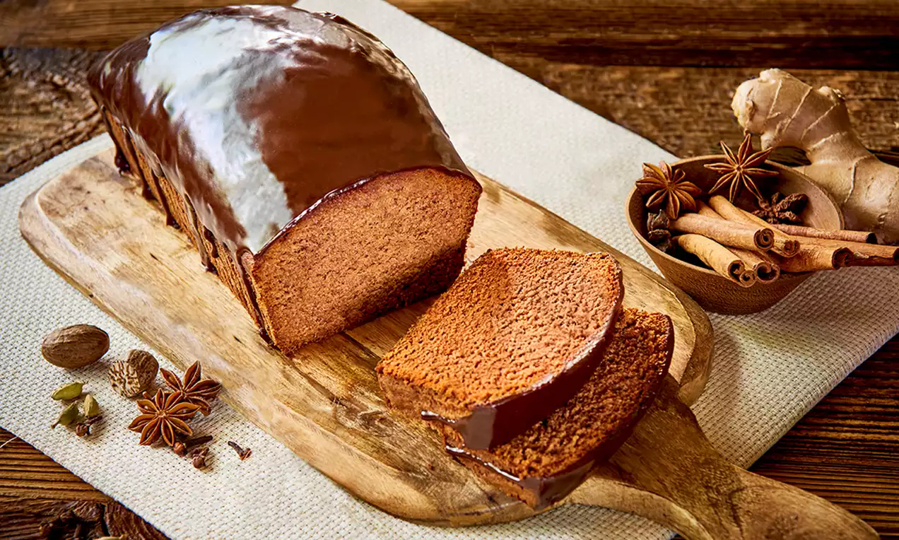 Domowy piernik w kształcie podłużnego bochenka, oblany czekoladową polewą, na drewnianej desce obok korzennych przypraw i imbiru
