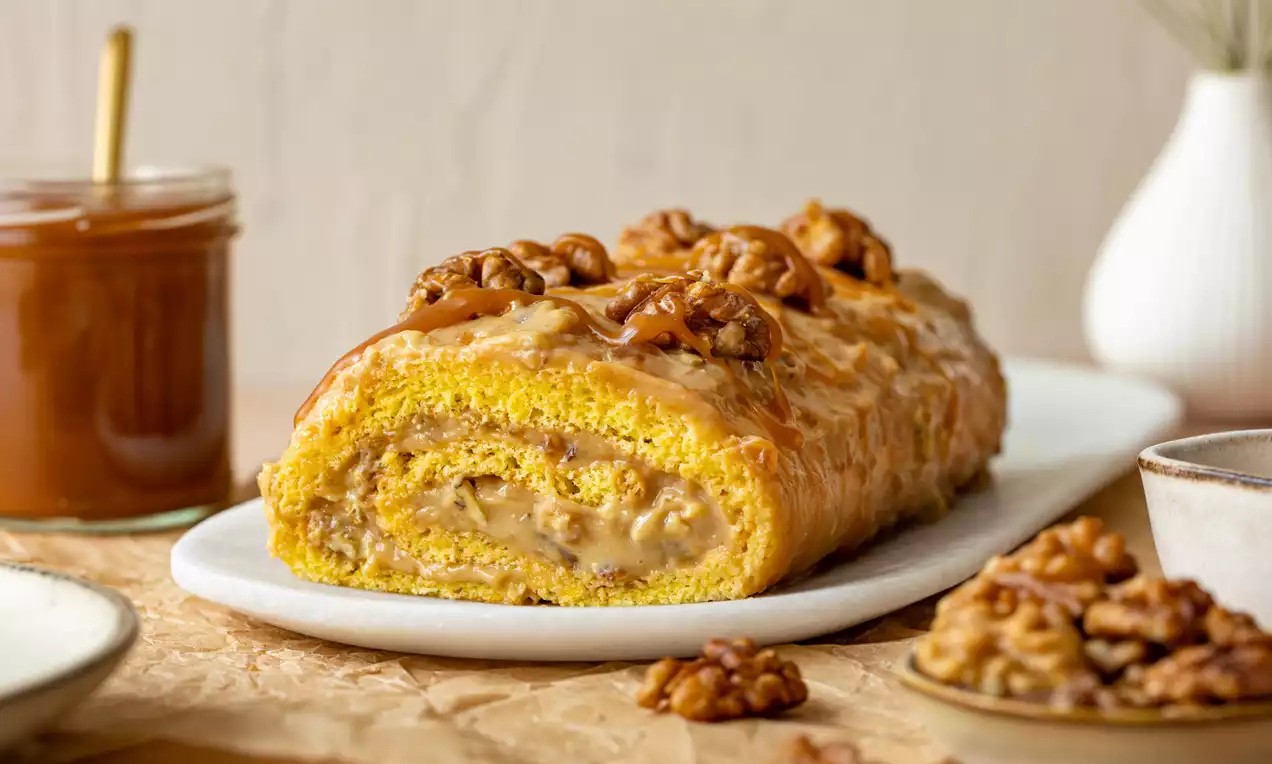 Zlatistá karamelová roláda plnená jemným orechovým krémom, obliata polevou, zdobená celými orechmi, položená na bielom podnose