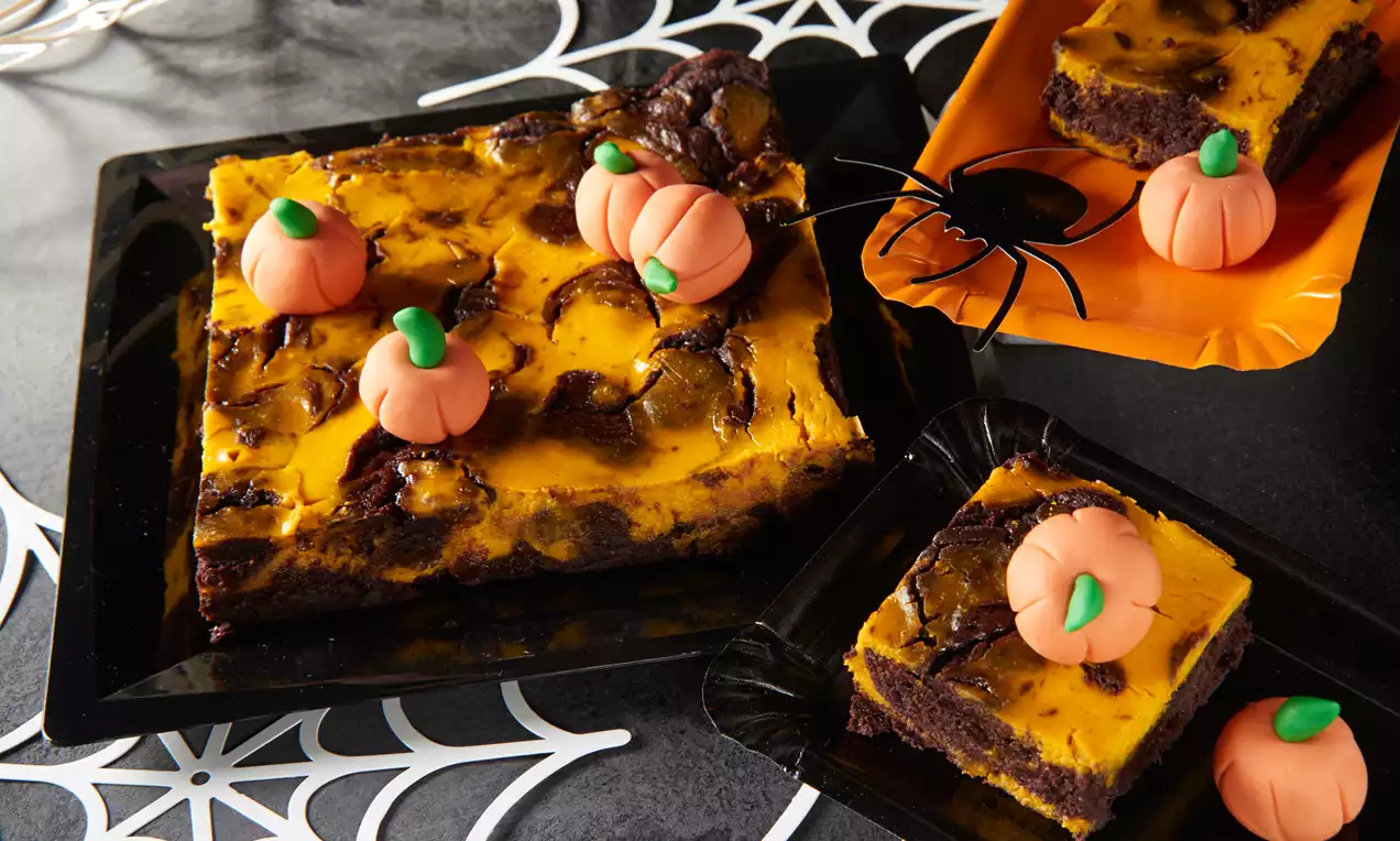 Kockasti brownie tamnosmeđe baze s narančastim kremastim preljevom, ukrašen modeliranim tikvicama na tamnim plastičnim tanjurima