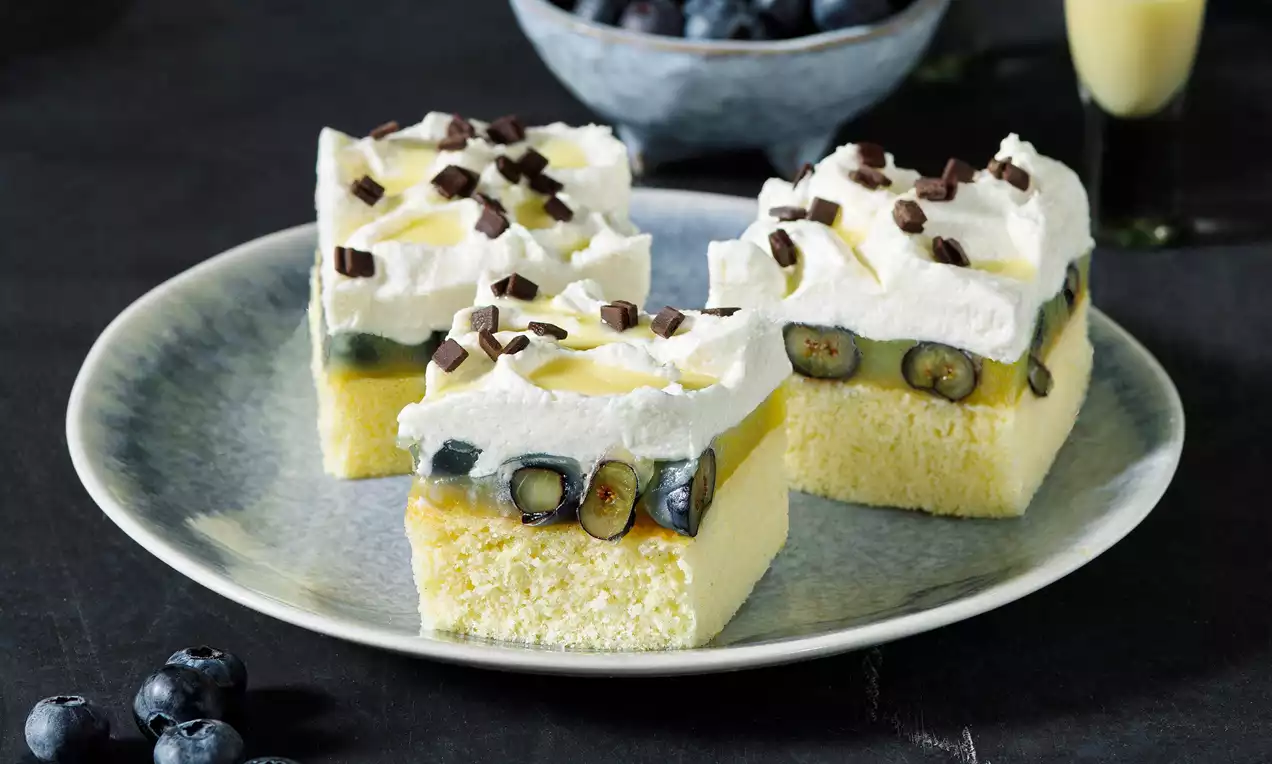 Kostky světlého piškotu s vrstvou borůvek, vaječného likéru a šlehačky, ozdobené čokoládovými hoblinkami na keramickém talíři