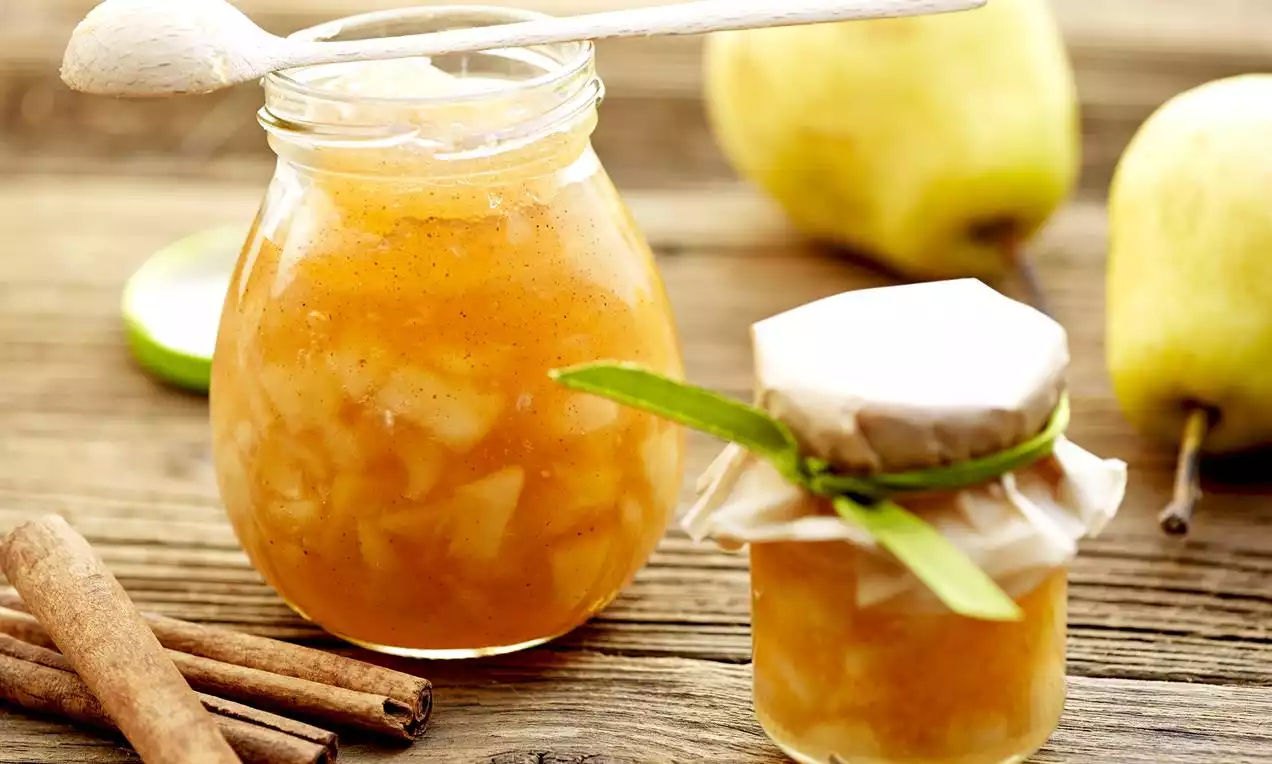 Marmellata dorata con pezzi di pera lucidi in un vasetto di vetro trasparente, decorata con bastoncini di cannella e pere mature sullo sfondo