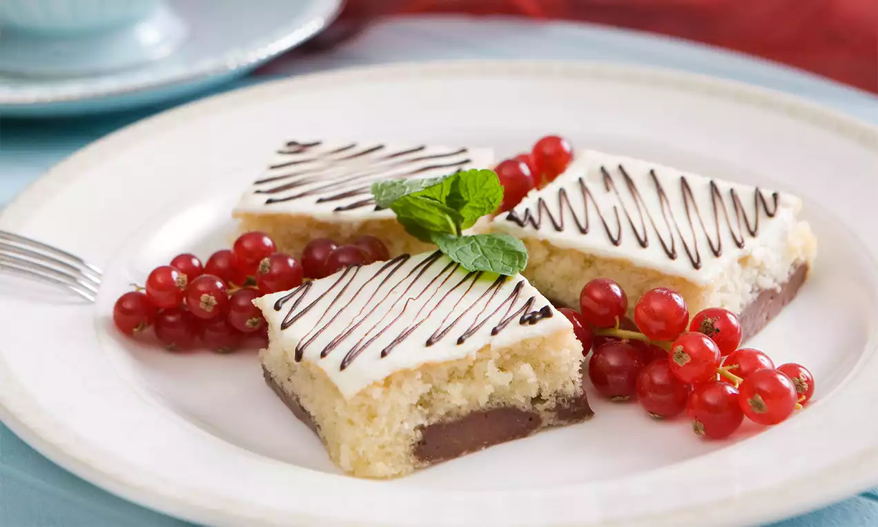 Kousky světlé mramorové bublaniny s čokoládovou vrstvou, bílovou polevou a čokoládovou dekorací, doplněné červeným rybízem a lístky máty