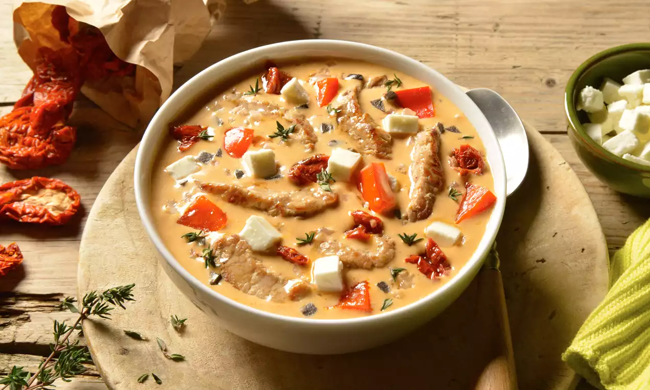 Gyrossuppe mit Gyrosstreifen in cremiger Brühe, roten Paprikastreifen, weißen Schafskäsewürfeln und grüner Petersilie in weißer Schale