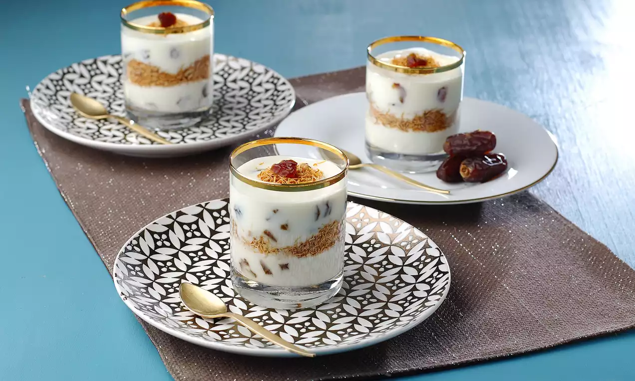 Küp bardaklarda katmanlı sunulan yoğurtlu tatlı, tel kadayıf ara katları ve hurma detaylarıyla desenli tabaklarda servis edilmiş