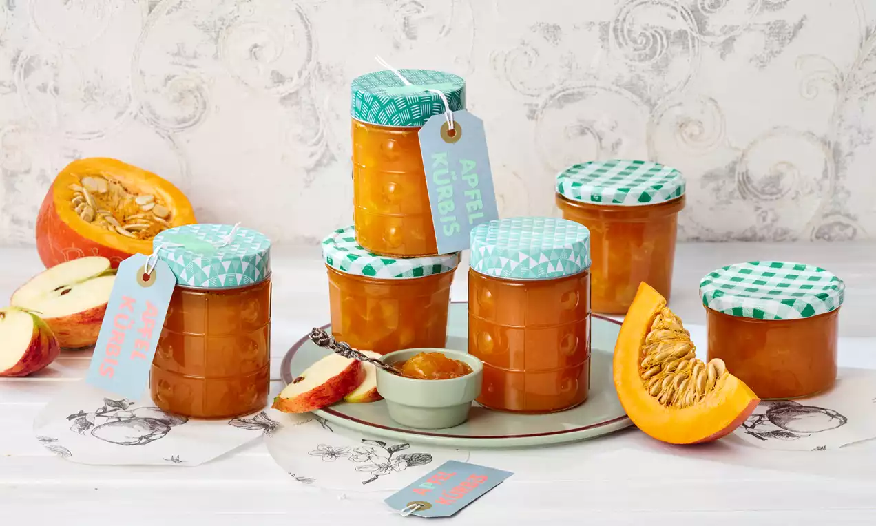 Gläser mit goldorangener Marmelade und grün gemusterten Deckeln, umgeben von Apfel- und Kürbisstücken auf hellem Untergrund