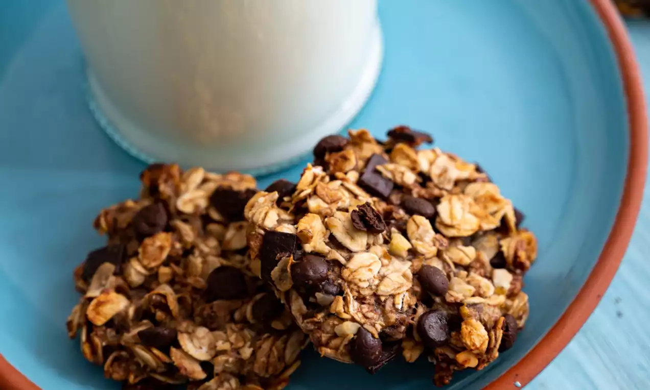 Zlatnosmeđi keksi s komadićima čokolade, neravnom rubnom teksturom, položeni na plavi tanjur uz visoku čašu mlijeka