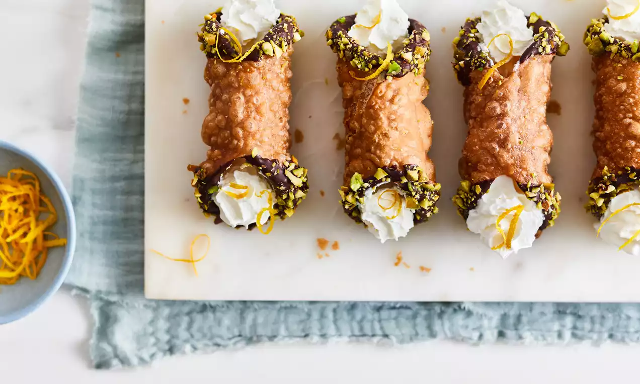 Four golden-brown vegan cannoli with crisp shells, chocolate-dipped edges, pistachio crumbs, whipped filling, and orange zest on marble