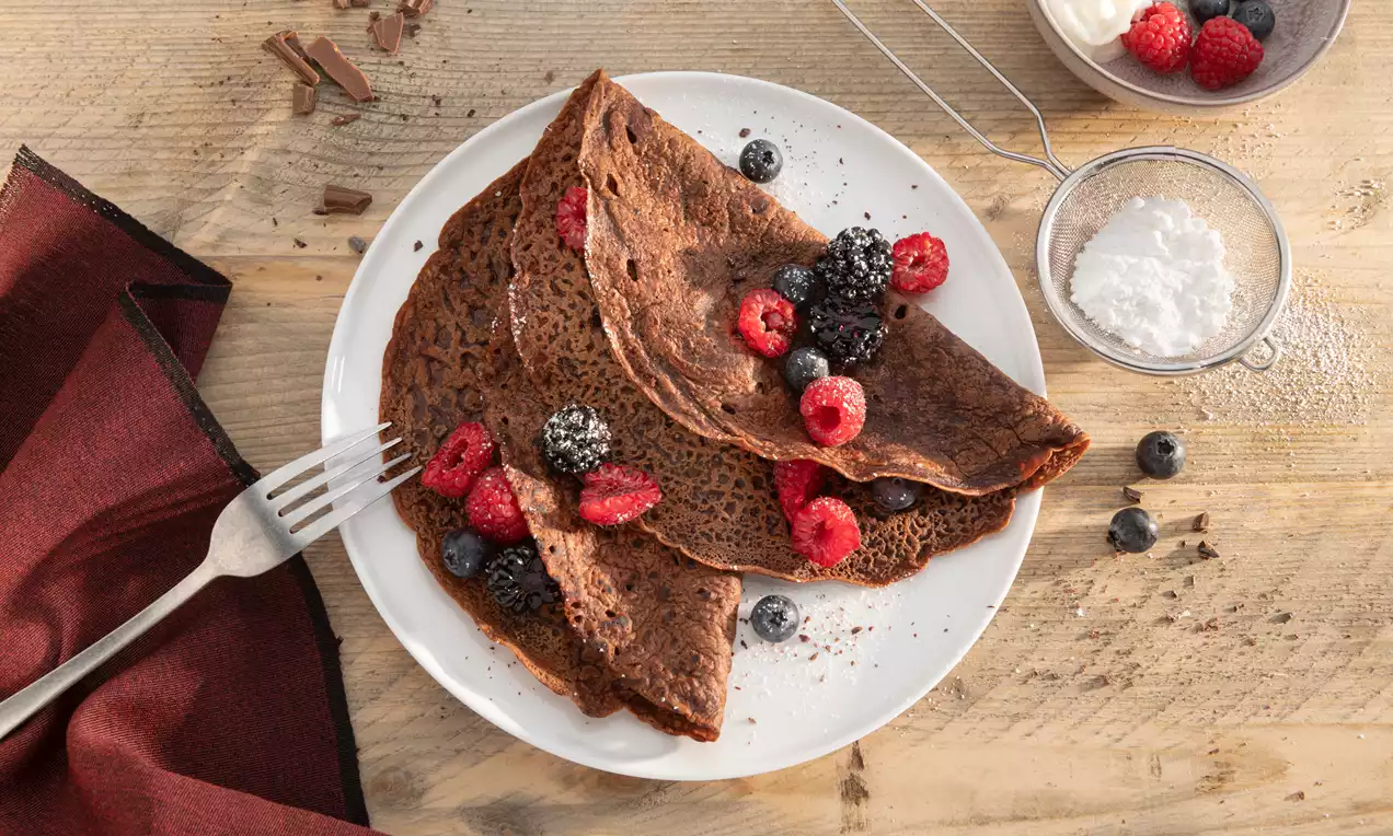 Dunne chocoladepannenkoeken op een wit bord, gegarneerd met sappige blauwe bessen en frambozen, omringd door poedersuiker en houten decor