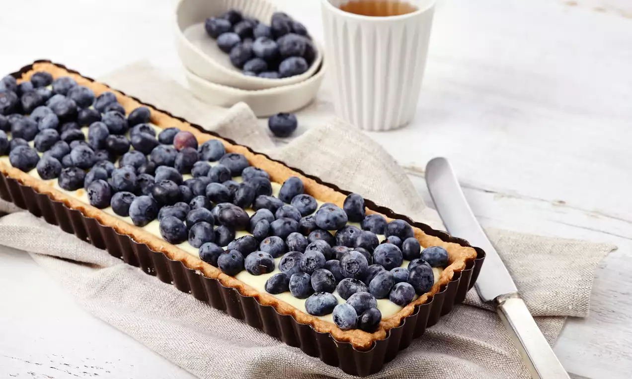 Pitkä suorakulmainen tartalettipohja täytetty kermaisella vanukkaalla, päällä tiiviisti aseteltuja mehukkaita mustikoita valkoisella pöydällä