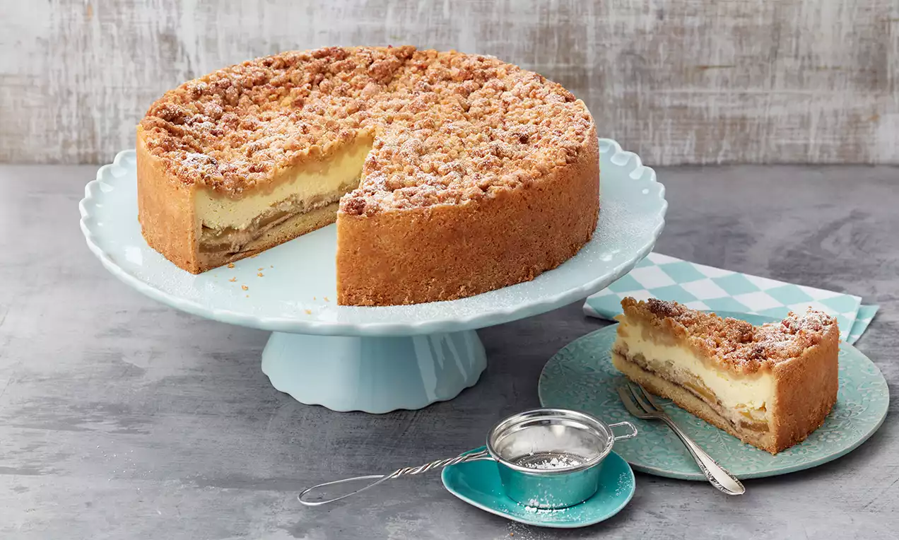 Tort de mere cu budincă cremoasă și strat crocant de crumble aurie, așezat pe un platou albastru, alături de o felie pe farfurie verde