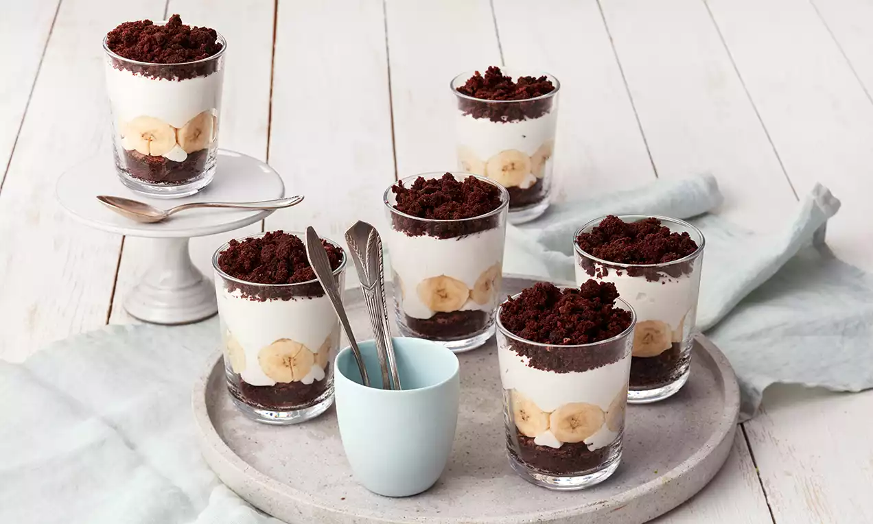 Schichtdessert im Glas mit hellem Sahnecremeband, Bananenscheiben und dunklem Kuchenkrümel-Topping auf grauem Teller mit Besteck im weißen Raum