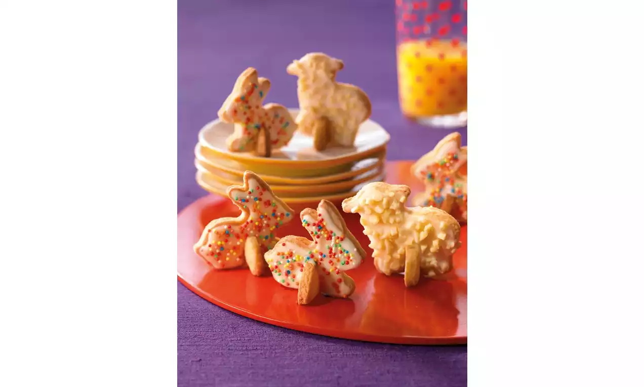 Citroenkoekjes in dierenvormen met glanzend gele glazuur en kleurige sprinkles op een rood bord, gestapelde borden en glas op paarse achtergrond