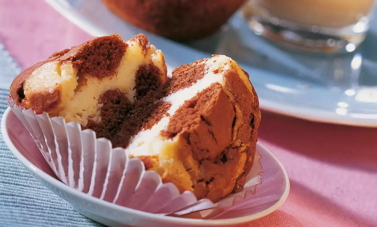 Muffin variegato al cacao e crema chiara, dalla superficie morbidamente screpolata, su un pirottino rosa e piatto bianco con sfondo colorato