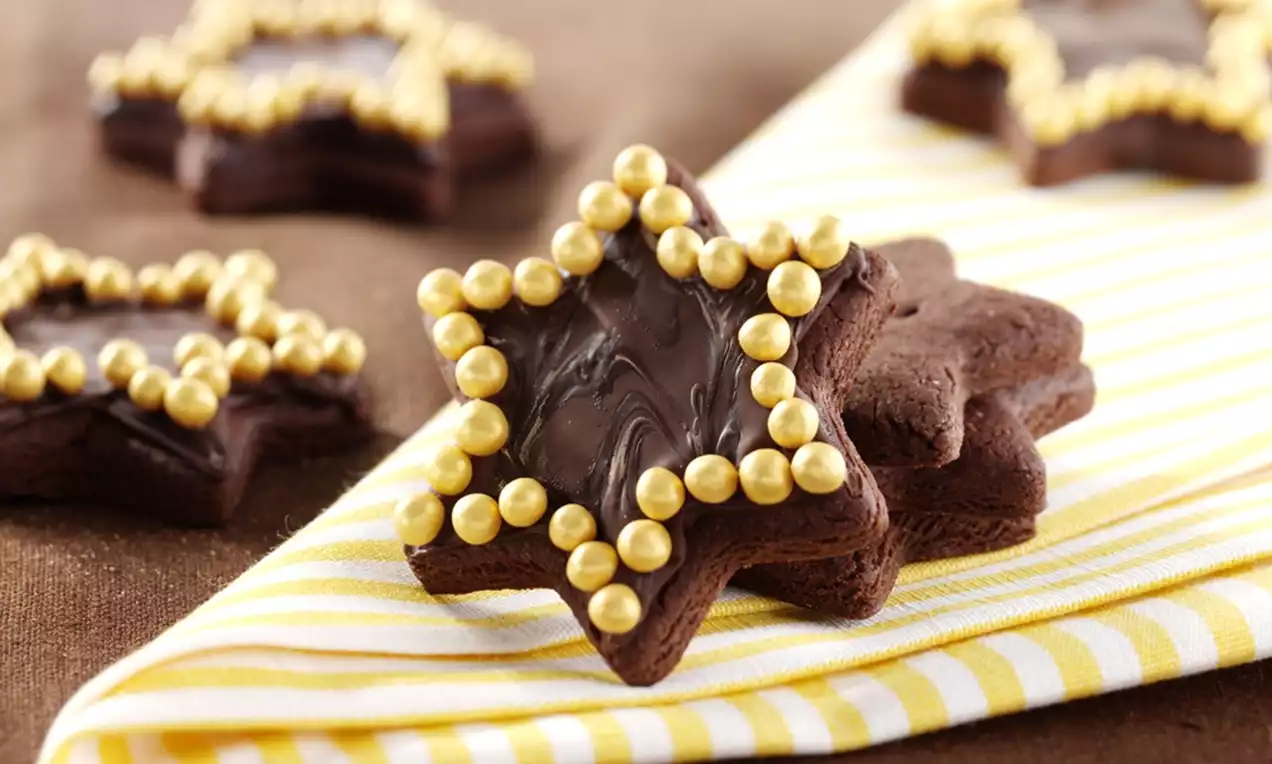 Dark chocolate star-shaped cookies with glossy swirled chocolate centers outlined in gold sugar pearls on a striped yellow cloth