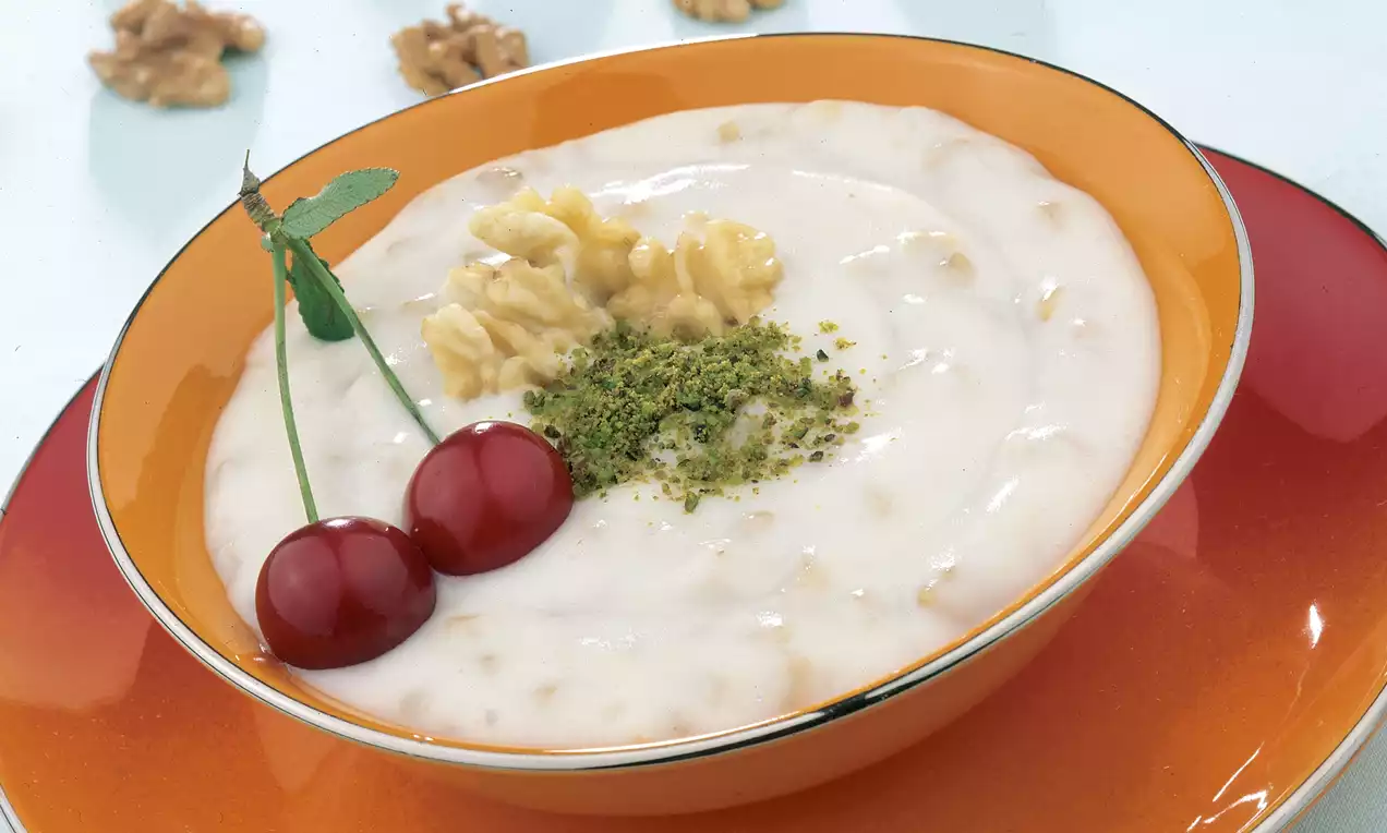 Portakal rengi geniş bir tabakta sunulan cevizli buğdaylı muhallebi, üzerine yeşil toz fıstık serpilmiş, iki kiraz ve ceviz parçaları ile süslenmiş