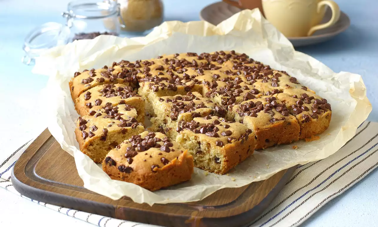Altın sarısı kek, kare dilimlenmiş, üzerinde bol çikolata parçaları, fırın kağıdı üzerinde ahşap bir sunum tahtasına yerleştirilmiş