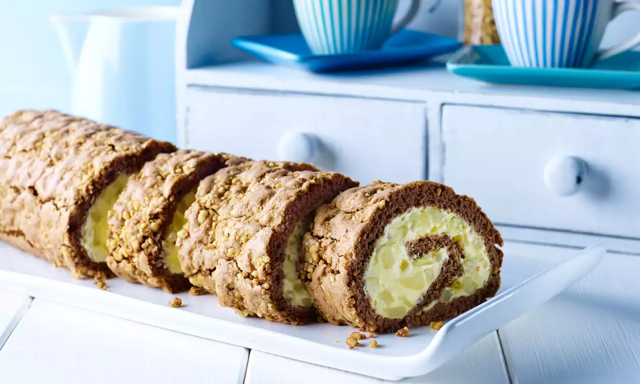 Gerollter Kuchen mit brauner Krokantkruste und heller Birnencremefüllung, in Scheiben auf einer weißen Platte angerichtet
