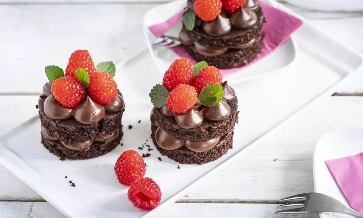 Mini round chocolate cakes layered with smooth chocolate cream, topped with fresh red raspberries and green mint leaves on a white plate